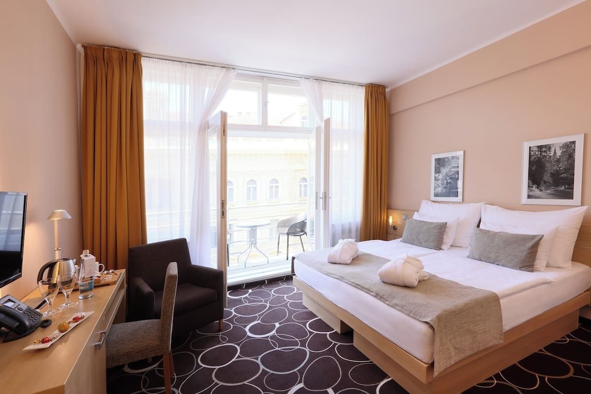 Habitación de hotel con cama, escritorio con teléfono y cafetera, una silla y vistas al exterior a través de un ventanal grande.