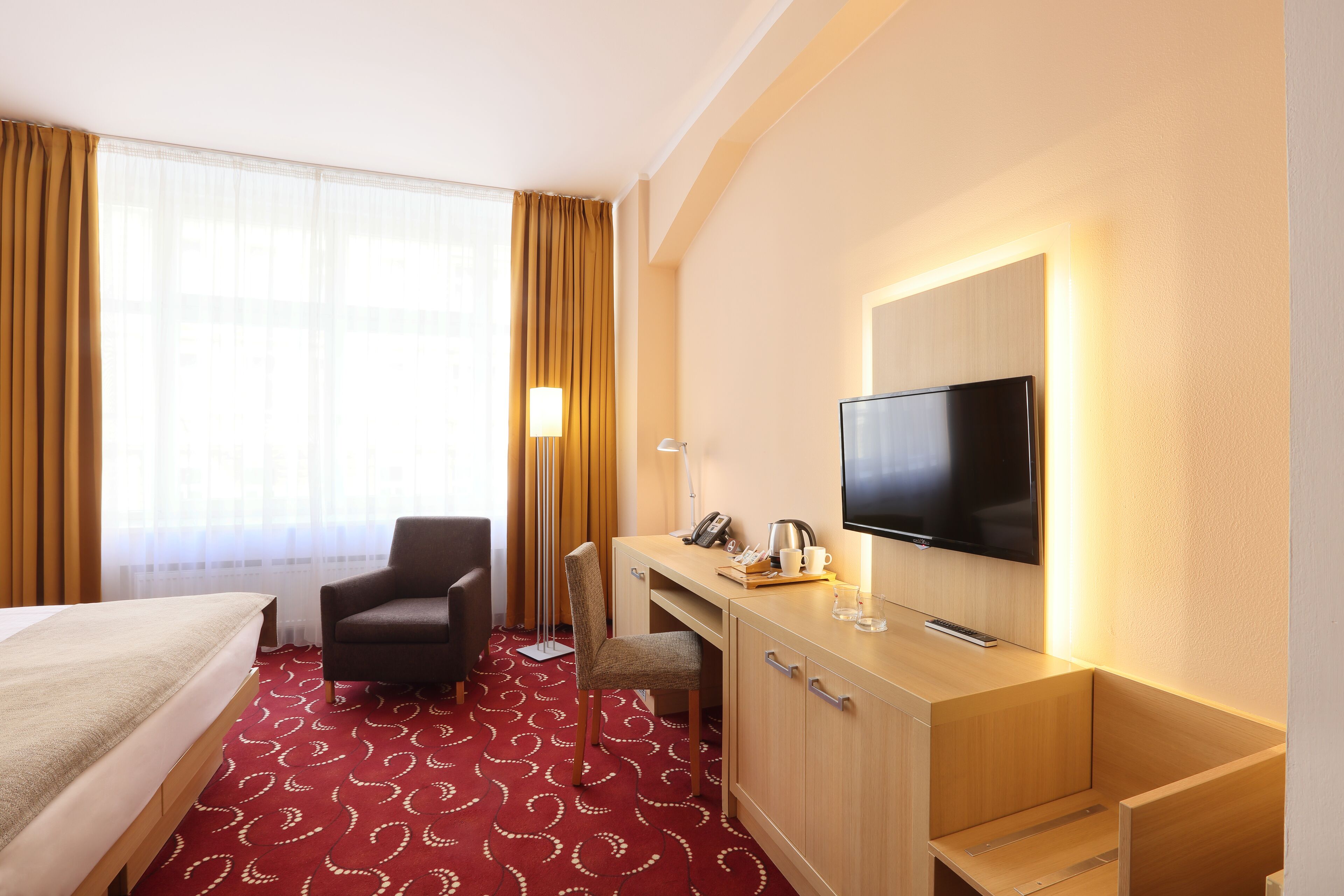 A hotel room with a bed, a desk with a TV, a chair, and a desk lamp.