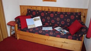 En soffa som kan fällas ut till en säng, tillverkad av trä, med en mönstrad kudde, en röd prydnadskudde och en liten pall.