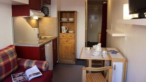 Une petite cuisine équipée comprenant un four à micro-ondes, un évier et des placards en bois. On y trouve une petite table à manger avec une chaise, ainsi qu’un canapé rouge avec un magazine.