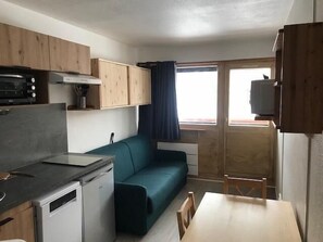 A compact kitchenette with a stove, microwave, and small refrigerator. A teal sofa and a wooden dining table with chairs are positioned near a window with curtains.