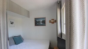 Un dormitorio pequeño con cama, televisor montado, un cuadro en la pared y una ventana con cortinas estampadas.