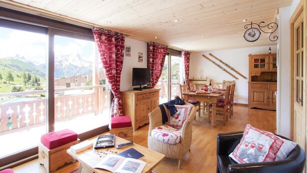 Ein gemütliches Zimmer mit Holzbalkendecke, einem Essbereich mit Tisch und Stühlen, einem Fernseher und Bergblick durch große Fenster.
