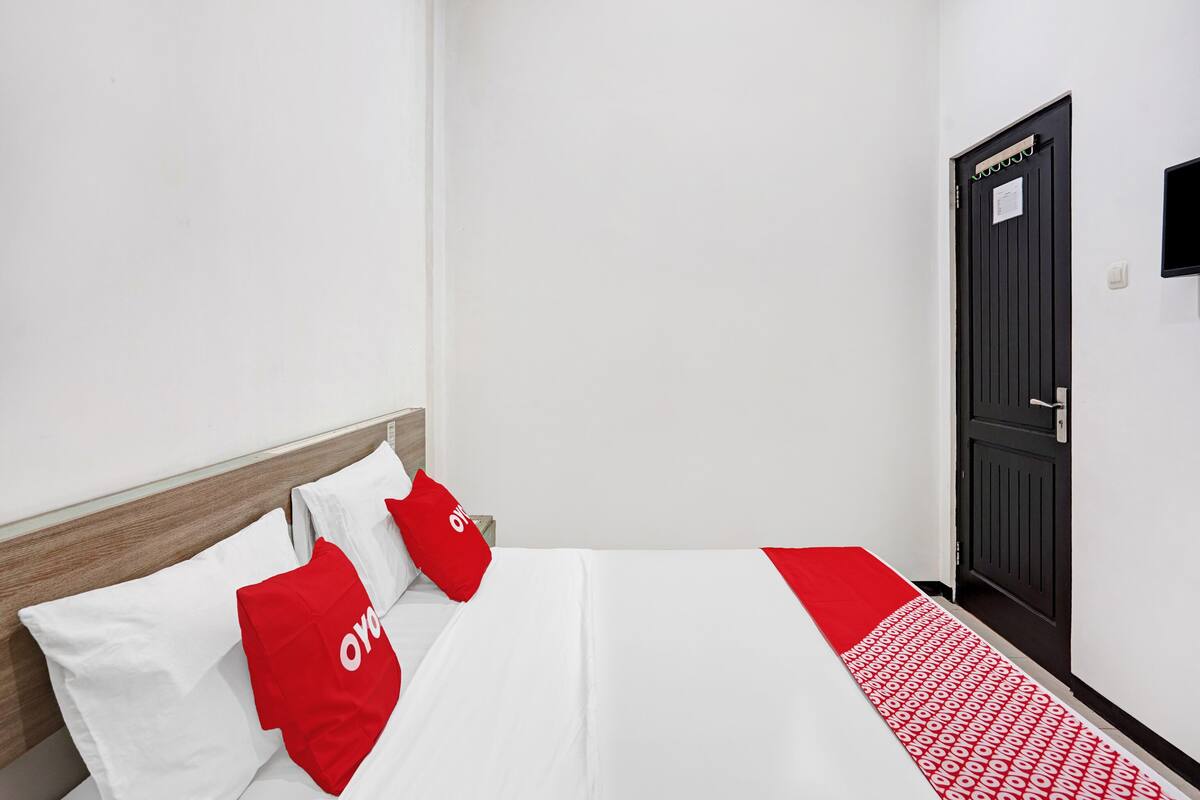Habitación de hotel con cama, paredes blancas, cabecero de madera y puerta negra.