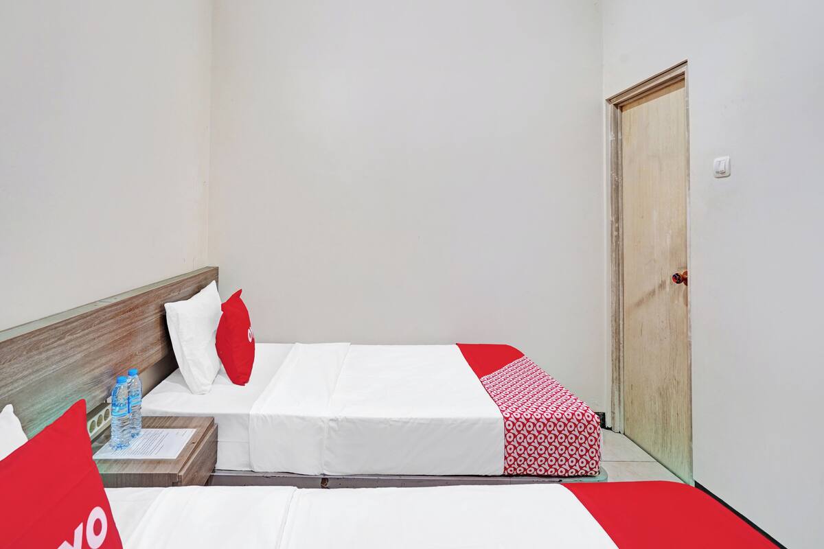 Habitación de hotel con dos camas, cabecera de madera, camino de cama rojo y blanco estampado, y una puerta con picaporte rojo.