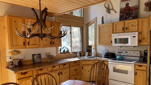 A wooden cabin kitchen with a chandelier, wooden cabinets, a dining table, and a poster on the wall. - Storybook Cabin - Private Stream, Incredible Canyon View, Starlink Internet (Bellvue)
