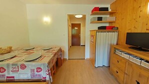Quarto compacto com uma mesa de jantar posta para quatro pessoas, uma televisão sobre um armário de madeira, e uma porta que dá para outro cômodo.