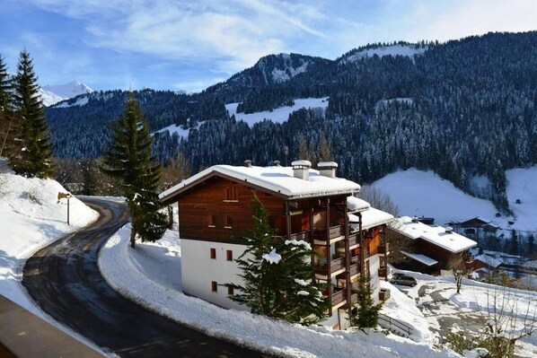 Un chalet en bois, avec un toit enneigé, un balcon et une vue sur une route sinueuse et des montagnes couvertes de neige.