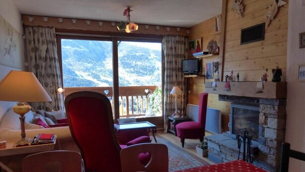 A cozy living room with a fireplace, a dining area with a table set for two, and a view of the mountains through the large window. - 200m from ski slopes, 4th floor, view mountain, balcony, fireplace or stove, tv, ski locker, 56m² (Méribel - Les Allues)