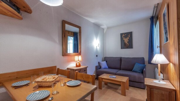 Un salon chaleureux avec une table à manger en bois, dressée pour deux personnes, un canapé et une petite table d’appoint avec une lampe.