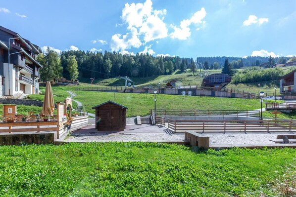 Ländliche Szenerie mit einer kleinen Holzhütte, einer gepflasterten Fläche mit Bänken und Blick auf einen bewaldeten Hügel im Hintergrund.