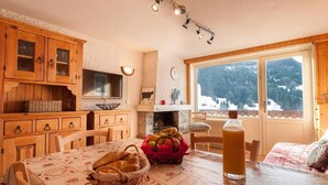 A cozy kitchen with wooden cabinets, a dining table with a red basket, and a large window with a snowy mountain view. - 3rd floor, balcony, fireplace or stove, tv, 63m², Le Grand Bornand (Le Grand-Bornand)