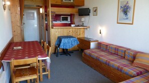 Een compacte kamer met een eettafel, stoelen, een bank, een kleine kitchenette en een televisie.