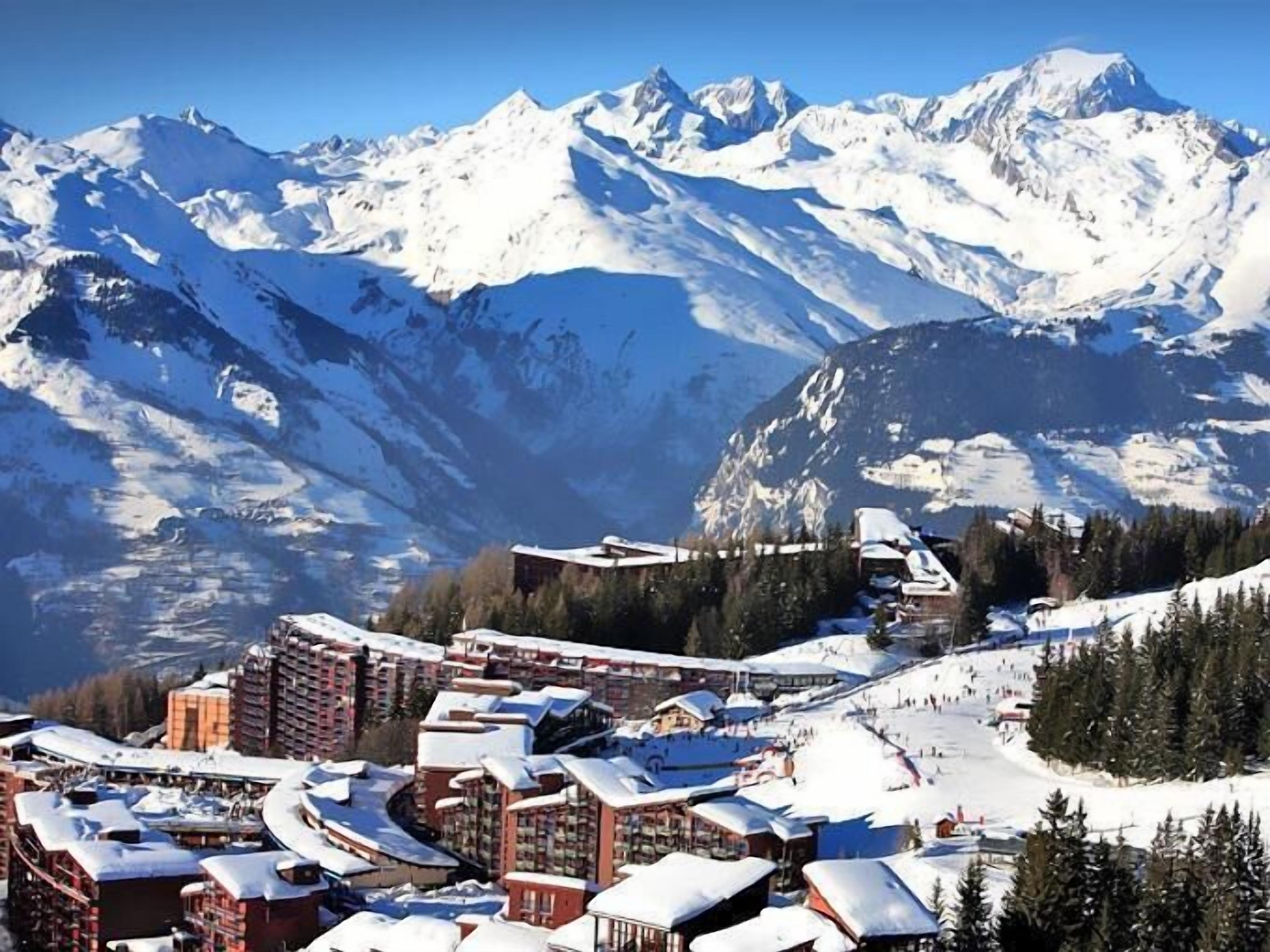 Ein schneebedecktes Bergresort mit mehreren Gebäuden, darunter ein Hotel, im Hintergrund ist eine Seilbahn zu sehen.