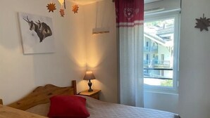 Une chambre à coucher avec une tête de lit en bois, un lit simple avec un oreiller rouge, une table de chevet avec une lampe, une fenêtre avec des rideaux et une œuvre d’art représentant un taureau, accrochée au mur.