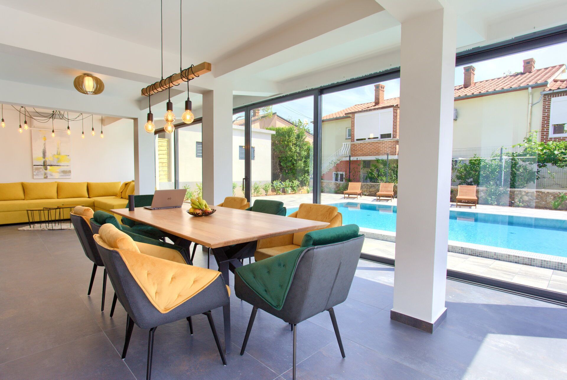 Un espace repas moderne avec vue sur la piscine, comprenant une table en bois, plusieurs chaises et un canapé jaune.