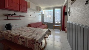 Une pièce confortable avec un lit, une petite table et un canapé rouge. Une fenêtre offre une vue sur les bâtiments à l’extérieur.