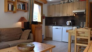 Un espacio habitable compacto con sofá, una mesa de madera, una zona de cocina con lavavajillas y una ventana con cortinas.