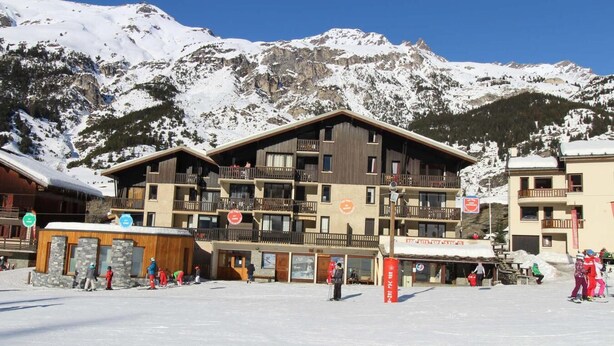 A multi-story building with balconies, ski rental facilities, and a ski slope in the foreground.