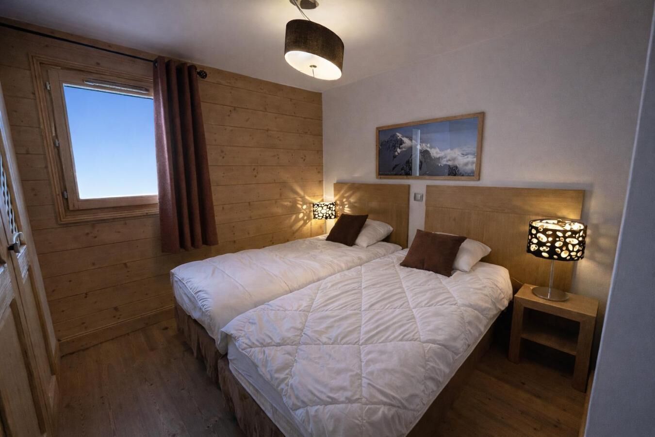 Une chambre avec deux lits, des murs en bois, une fenêtre avec des rideaux, un luminaire au plafond, une table de chevet avec une lampe et un tableau encadré au mur.