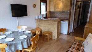 A dining area with a round table set for four, wooden chairs, a kitchen with wooden cabinets, a built-in dishwasher, and a flat-screen TV mounted on the wall.