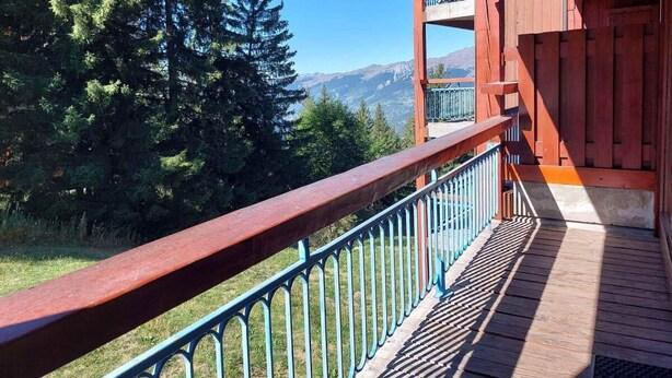 Un balcon en bois, doté d’une rambarde en métal, qui offre une vue sur une zone boisée et des montagnes.