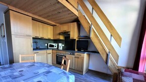 A compact kitchen with wooden cabinets, a microwave, and a stove. There's a dining table with a blue tablecloth and a chair. A wooden staircase leads to an upper level. - 150m from ski slopes, view mountain, balcony, tv, 35m², Les Menuires (Les Menuires)