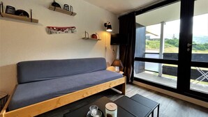 A modern room with a wooden sofa, a small table with a candle and a cup, and a view of greenery through large windows.