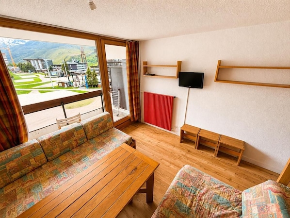 Ein Zimmer mit großem Fenster, Holzmöbeln, einem roten Heizkörper und einem an der Wand befestigten Fernseher.