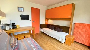 Ein Zimmer mit einem Bett, einem Tisch, einem Sessel, einem kleinen Hocker, einem Fernseher und einem roten Heizkörper.