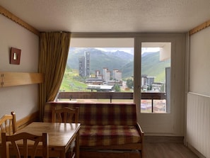 A room with a dining table and chairs, a bench, a window with a view of buildings and mountains.
