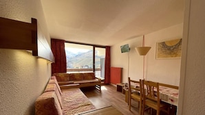 A compact room with a sofa set, a dining table with chairs, a small table, and a view of mountains through a large window. - 10m from ski slopes, 7th floor, balcony, tv, 36m², Les Menuires (Les Menuires)