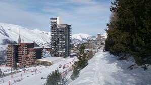 Una pista de esquí cubierta de nieve con edificios al fondo, incluyendo una estructura moderna y varios edificios más pequeños.