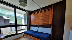 A bedroom with a wooden bed, a window with a view of hills, and a small table. - 300m from resort center, 3rd floor, view mountain, balcony, tv, ski locker, 20m², Les Menuires (Les Menuires)