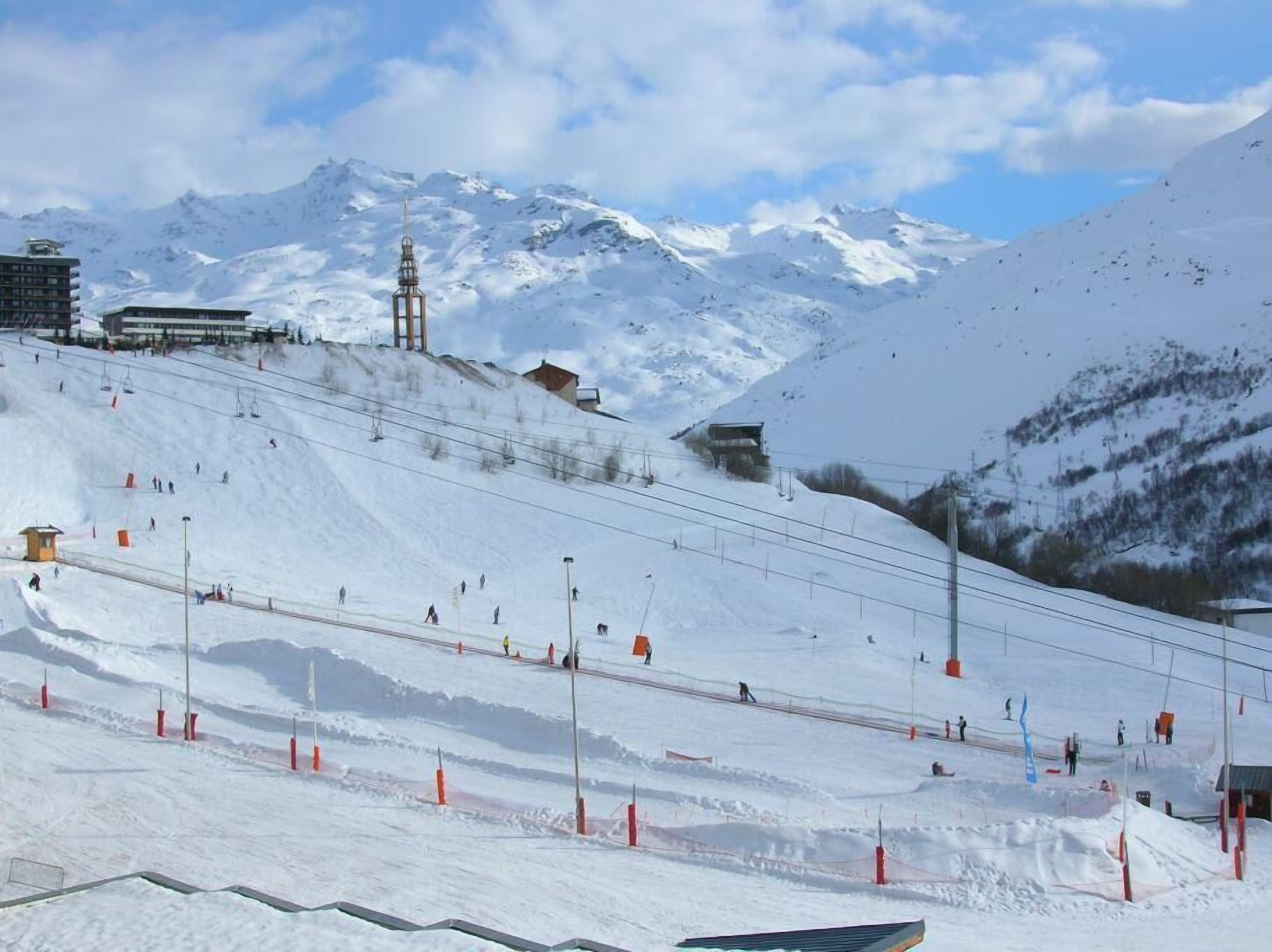 Ein verschneiter Skipisten mit Seilbahnen, einem Berg im Hintergrund und Gebäuden am Fuß.