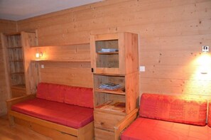 Een houten kamer met twee houten bedden met rode beddengoed, een houten kast met planken, en een wandlamp.