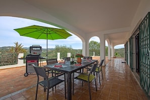 Um pátio externo com mesa posta para refeições, churrasqueira e guarda-sol verde.