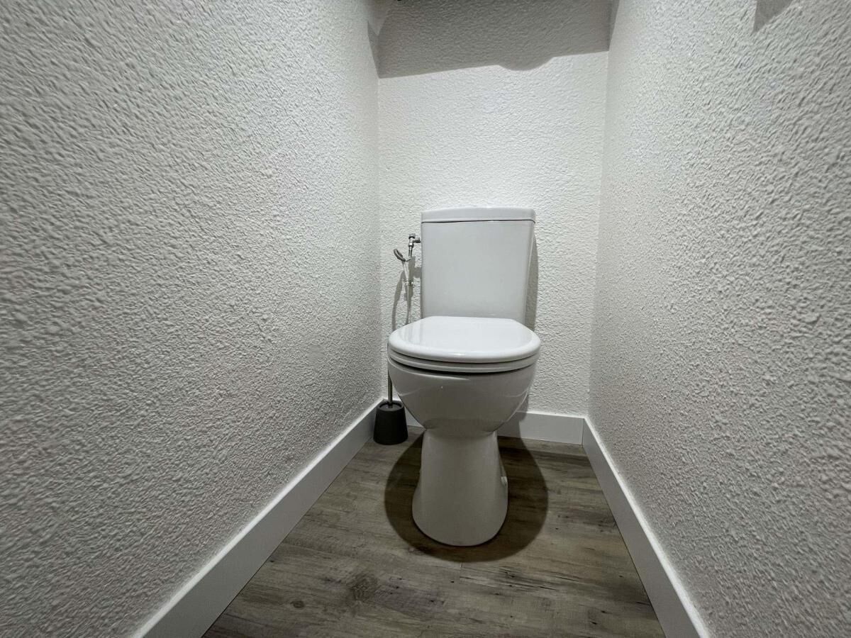 A small, narrow bathroom with a white toilet, a wooden floor, and textured walls.
