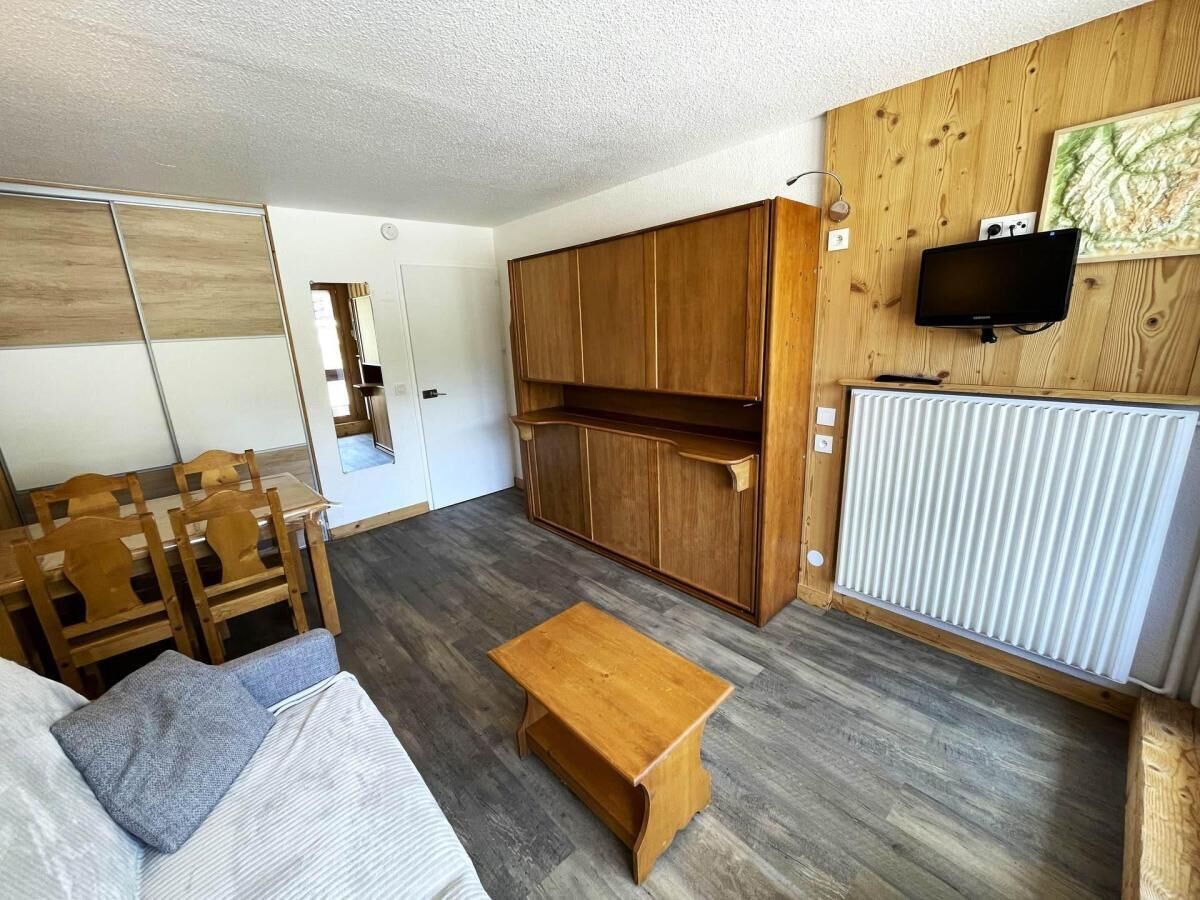 Ein kompakter Wohnbereich mit einer Couch, einem Holzesstisch und einem kleinen Beistelltisch. Ein Fernseher ist an der Wand montiert, und ein Heizkörper ist zu sehen.