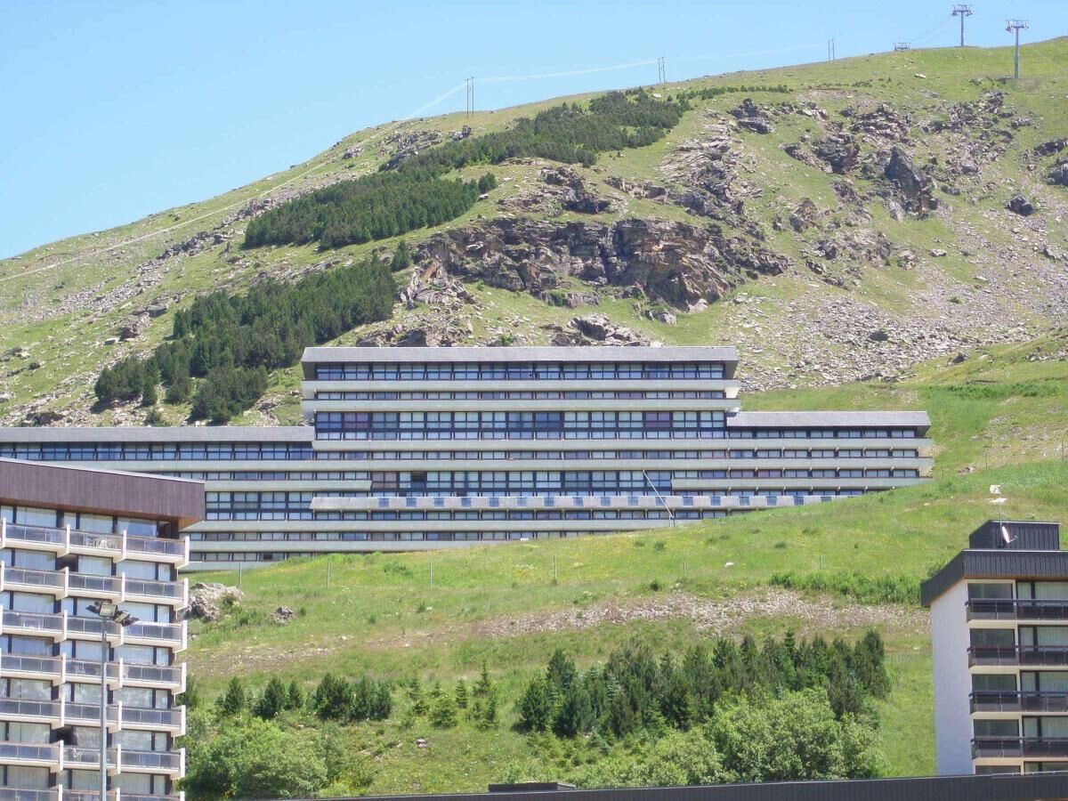 A modern multi-story building with a flat roof and a series of balconies, situated on a hillside with greenery and rocky terrain in the background.