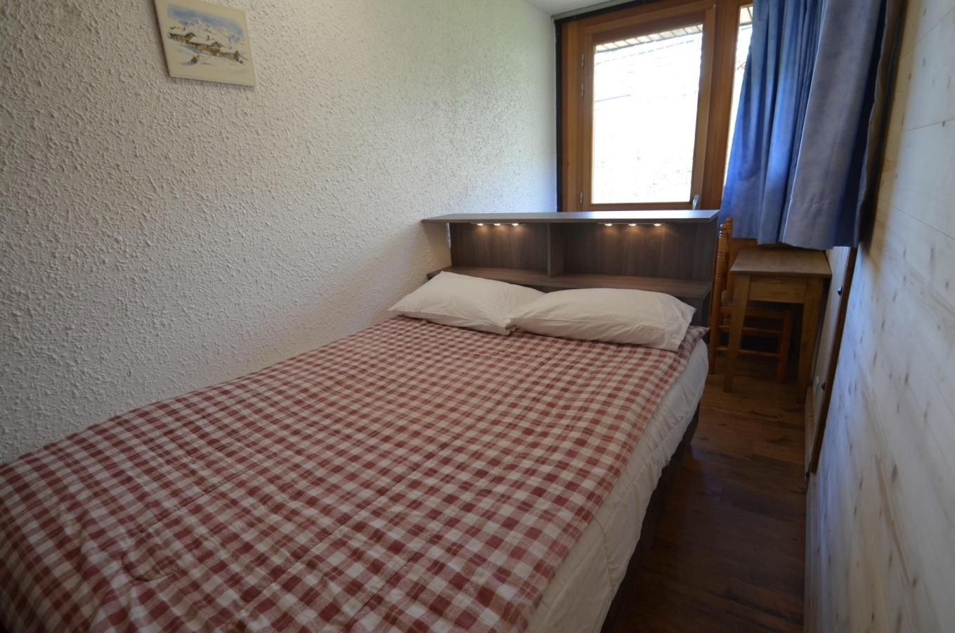 A single bed with a checkered bedspread in a room with a wooden headboard, a small window with curtains, and a picture on the wall.