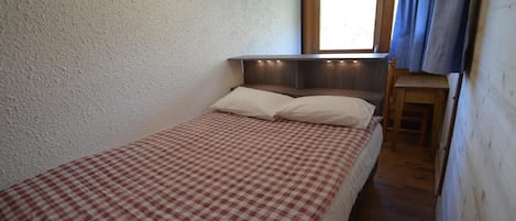 En säng med en rutig sängkappa i ett rum med en sänggavel i trä, ett litet fönster med gardiner och en tavla på väggen.