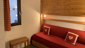 Una stanza piccola e accogliente con un divano rosso, due cuscini a forma di cuore, una testiera in legno, un tavolino in legno e una finestra con tenda a quadri.