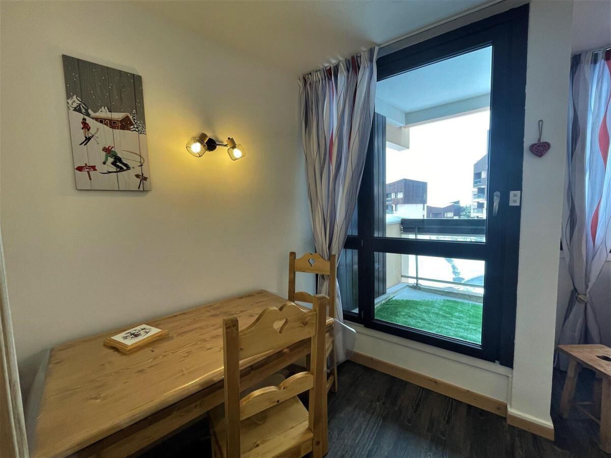 A room with a wooden table and chairs, a painting on the wall, a window with curtains, and a view of buildings outside.