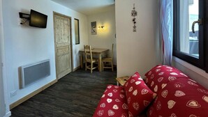 Una stanza compatta con un letto, un tavolino e una sedia. Si vede una finestra con tende, una TV a parete e un lampadario.