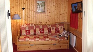 Une chaleureuse cabane en bois avec un canapé matelassé, un tableau encadré, une lampe et une petite table recouverte d’une nappe rouge.