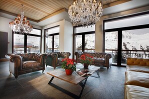 A spacious lounge with leather sofas, a wooden coffee table, and a large chandelier. Large windows offer views of a snowy mountain. - Wifi, ski-in/ski-out & foot of the slopes, fitness, swimming pool, sauna, hammam, balcony, tv (Les Arcs)