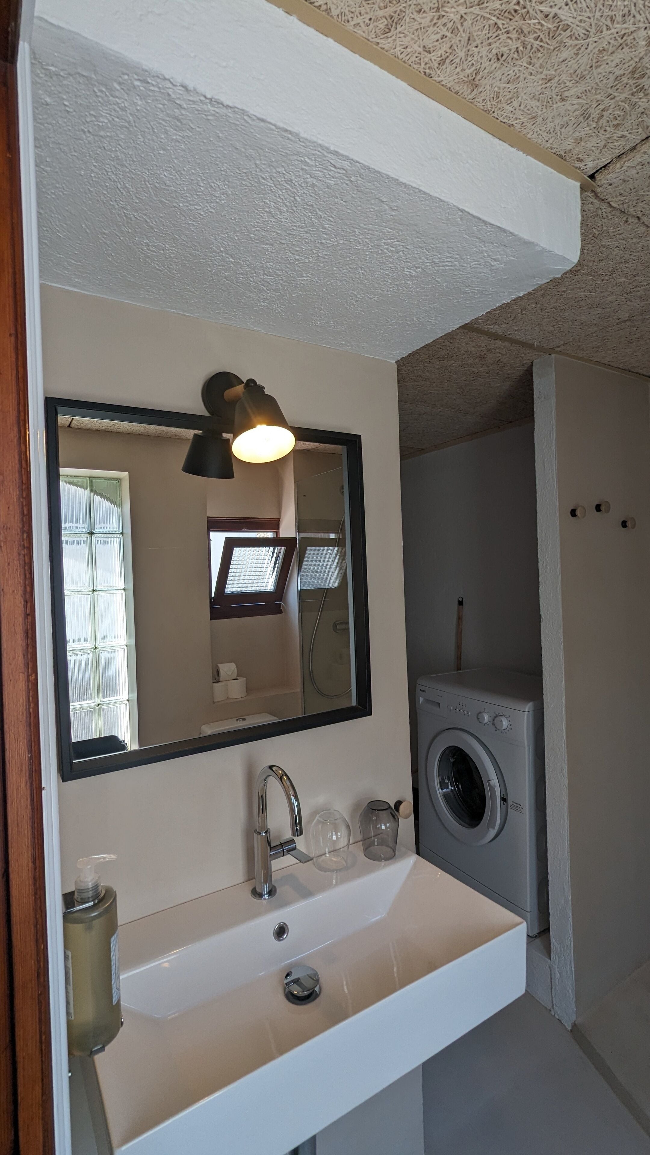 A bathroom with a white sink, a mirror, a black pendant light, a window, and a washing machine.