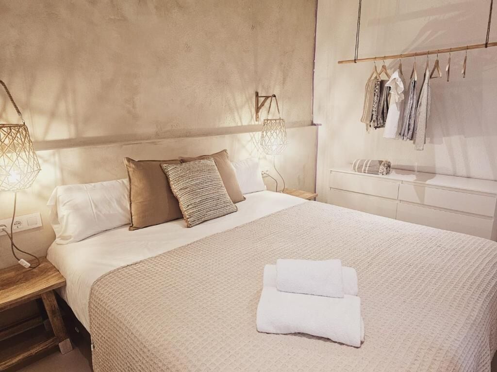 A neatly made bed with a folded towel, a bedside table with a lamp, and a clothing rack with garments.