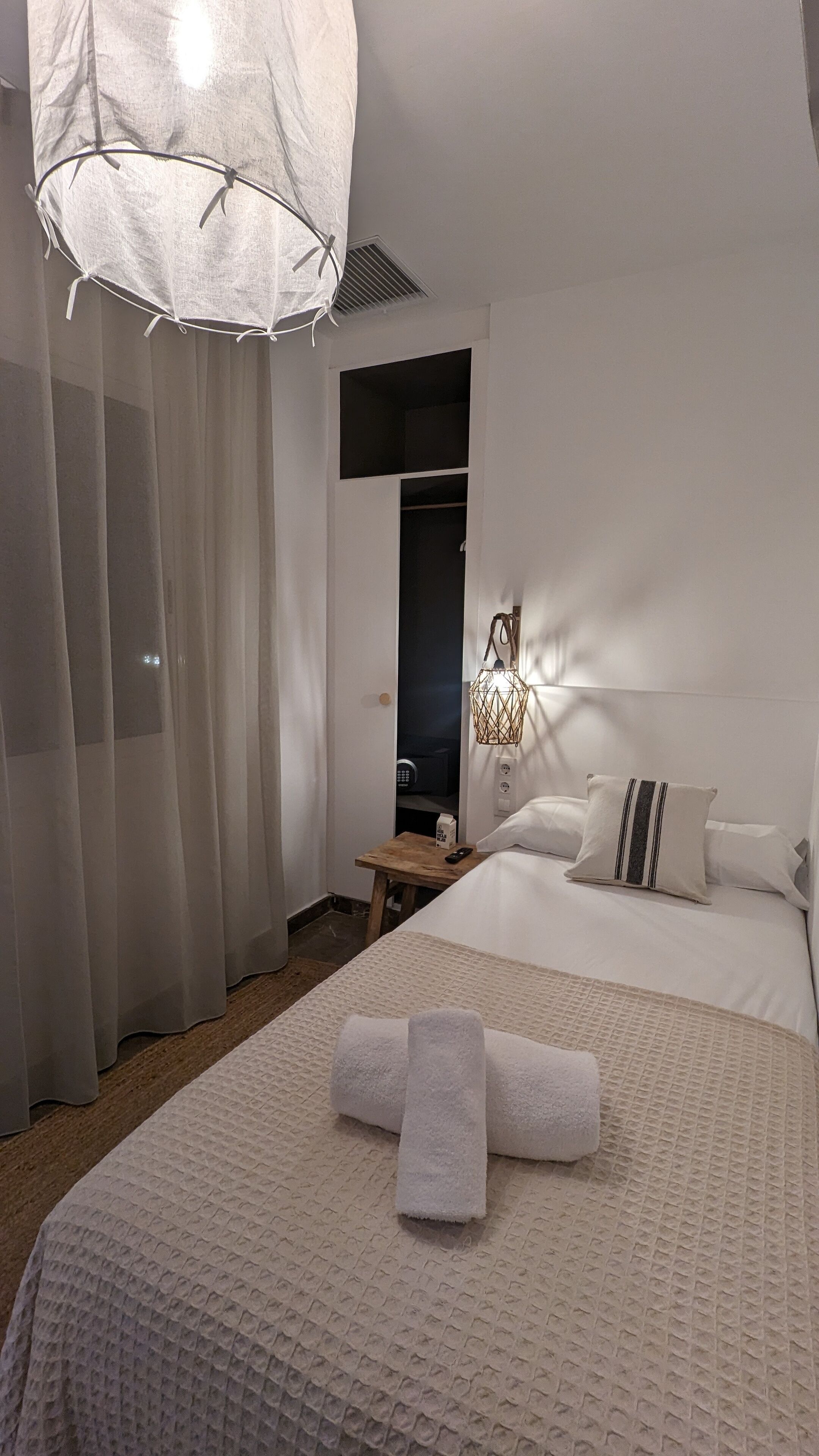 A bedroom with a bed, a bedside table, a nightstand, a hanging light fixture, and a closet.